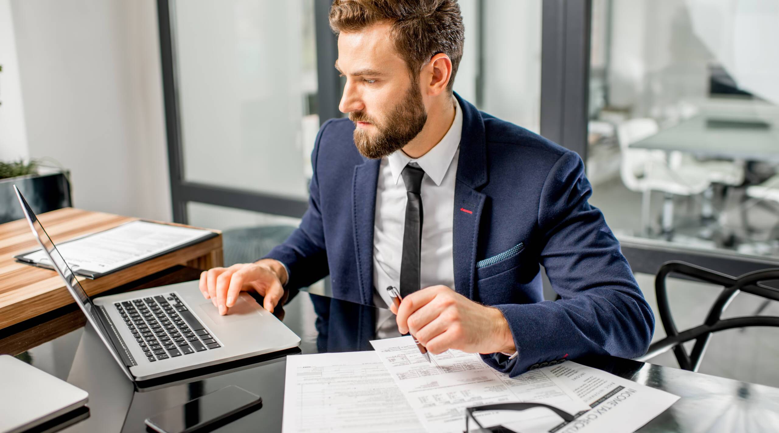 Tax manager working at the office Handsome tax manager dressed in the suit working with documents and laptop at the modern office interior